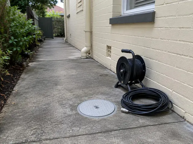 Flush round inspection cover on a concrete side path beside a coiled drain camera reel and brick wall in Hurlstone Park