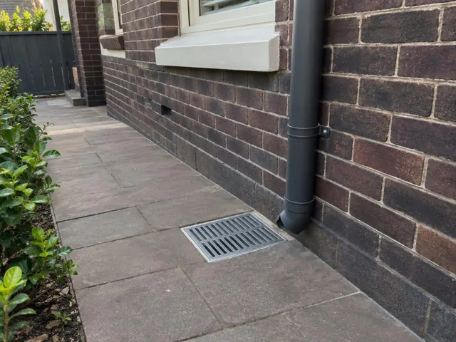 Outdoor drain gully with grate set into paved path beside a brick house in Hurlstone Park