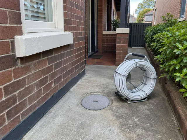 Capped access point in a concrete path beside a brick house and garden bed in Ashfield
